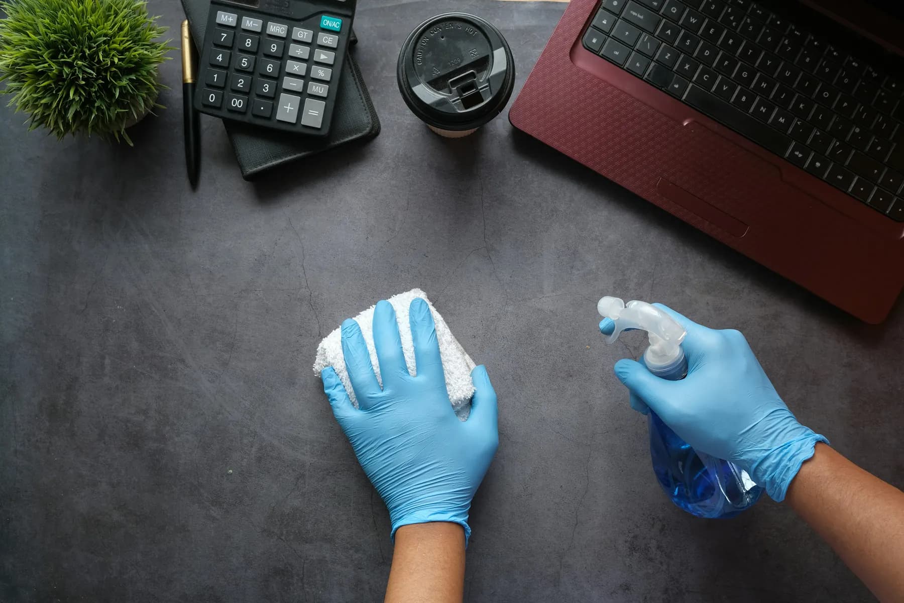 Gloved hands disinfecting a desk surface beside a laptop and calculator, an example of cleaning for gulf coast facilities