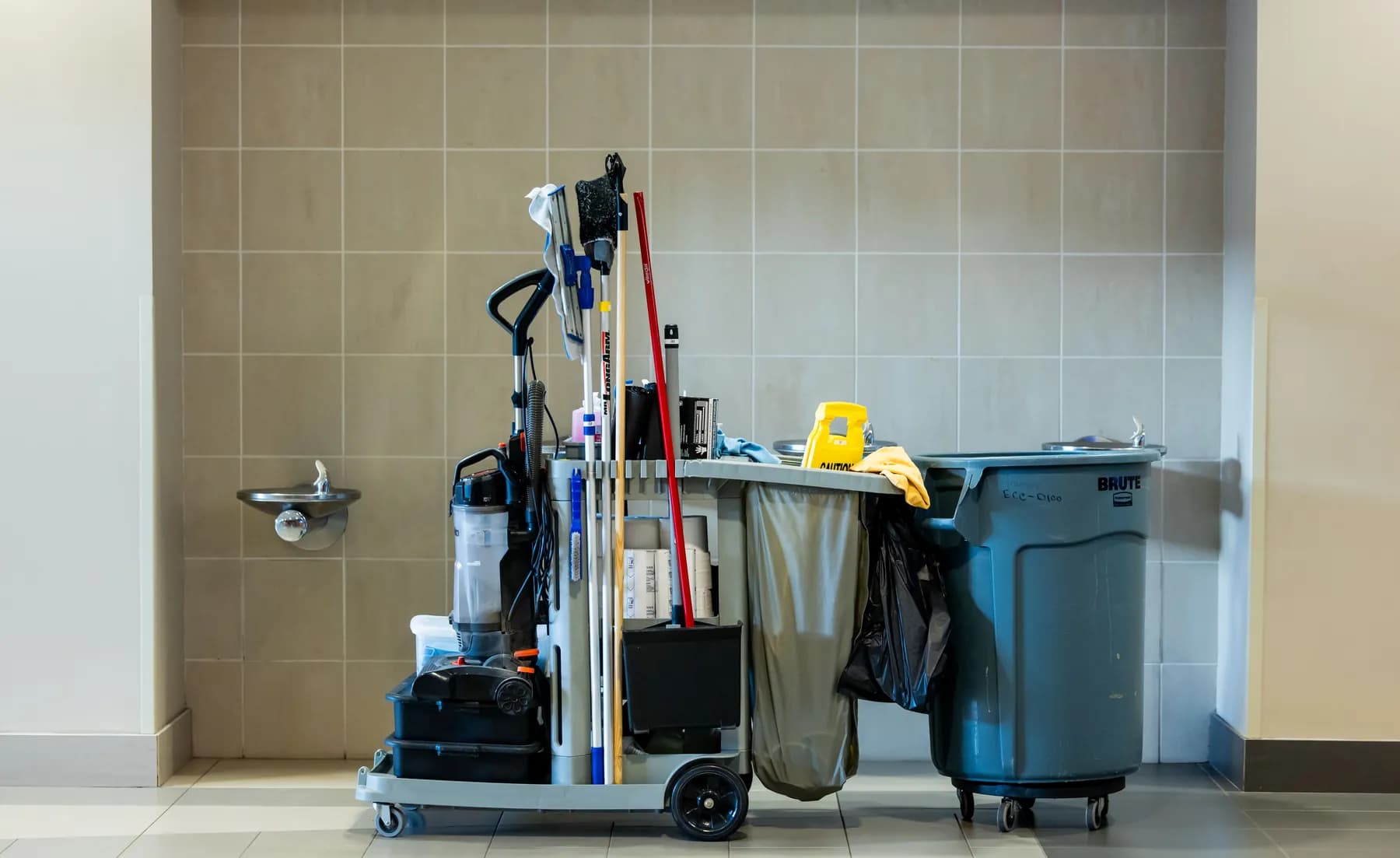 A stocked janitorial cart and waste bin parked against a tiled corridor wall, an example of commercial cleaning services