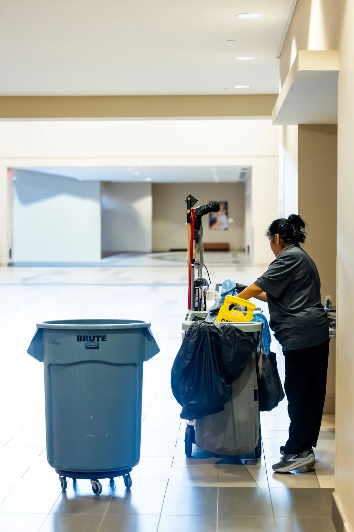 A janitorial cart and waste bin beside a worker in a large tiled building lobby, an example of janitorial services